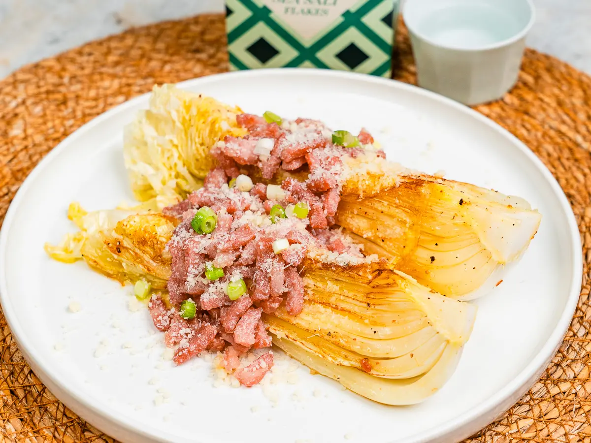 scallions being sprinkled over plated cabbage steaks with cheese and bacon