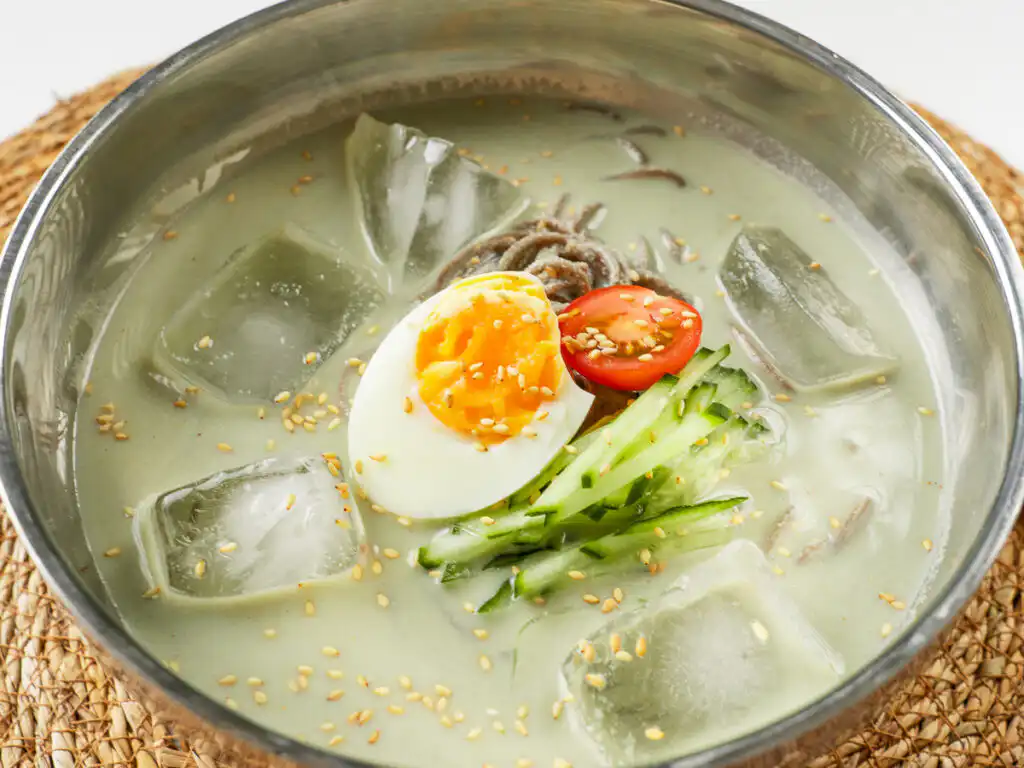 Close-up of matcha kongguksu broth swirling around cold buckwheat noodles in a stainless steel bowl
