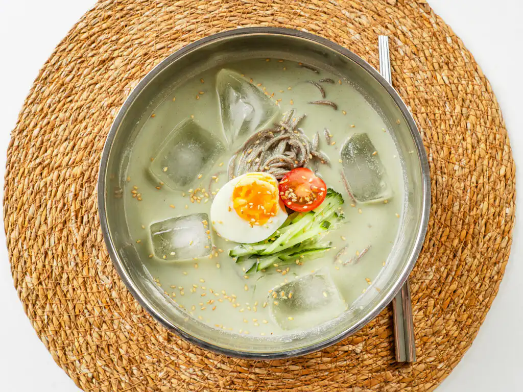 Overhead shot of matcha kongguksu with ice cubes floating in the green broth, metal chopsticks on the side