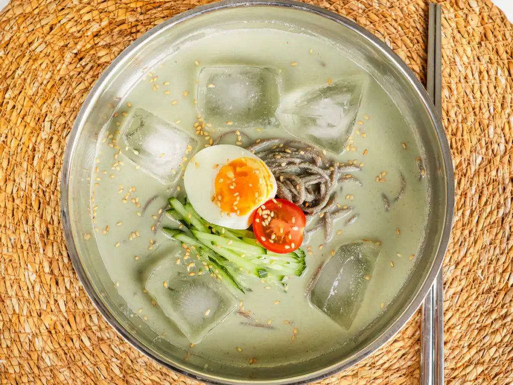 Overhead shot of matcha kongguksu with ice cubes floating in the green broth, metal chopsticks on the side