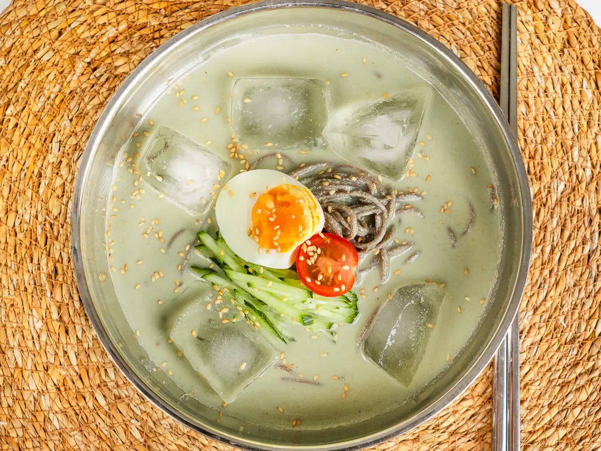 Overhead shot of matcha kongguksu with ice cubes floating in the green broth, metal chopsticks on the side