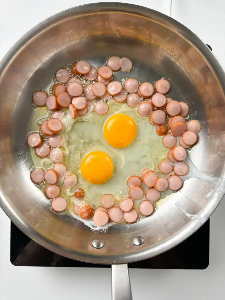 Two eggs cracked directly into pan alongside golden sausage for sausage and rice recipes Korean bowl