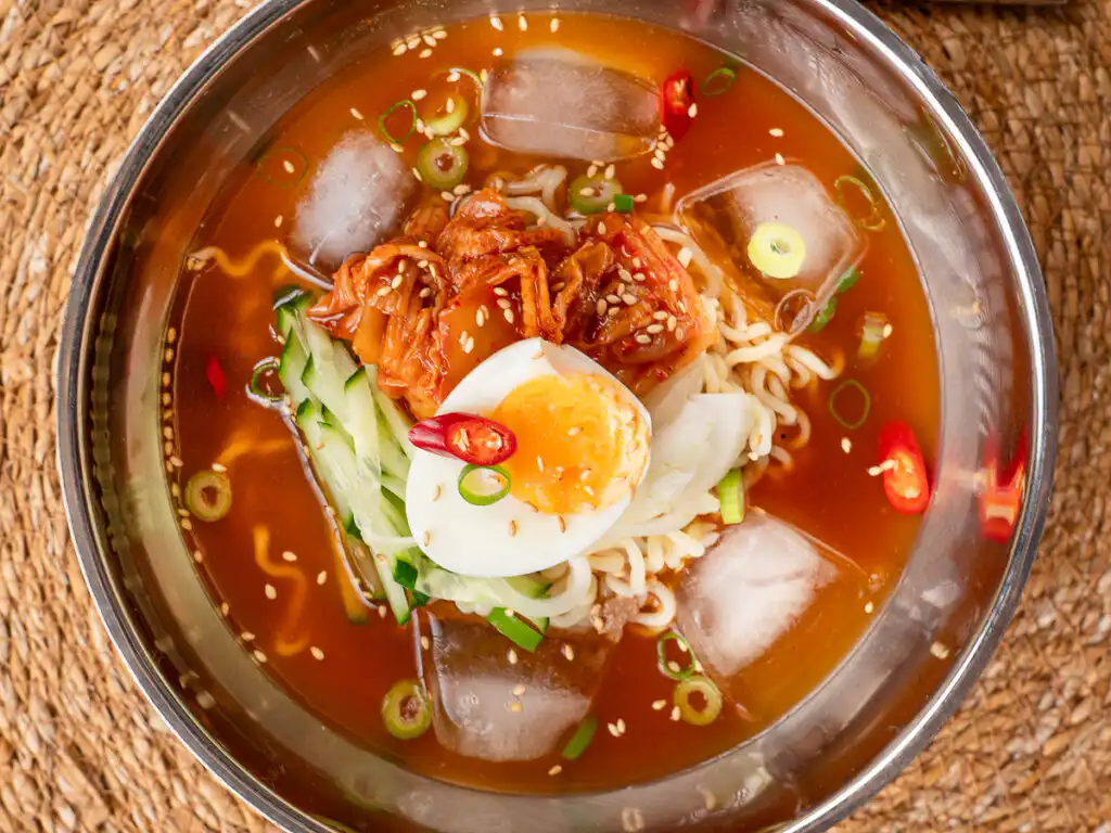 A bowl of cold ramyeon topped with kimchi, cucumber, egg, and sesame seeds