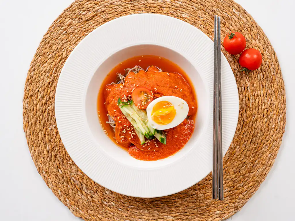 Tomato Gochujang Bibim Guksu - Summer Spicy Cold Noodles gochujang bibim guksu served on a white plate with chopsticks and side dishes