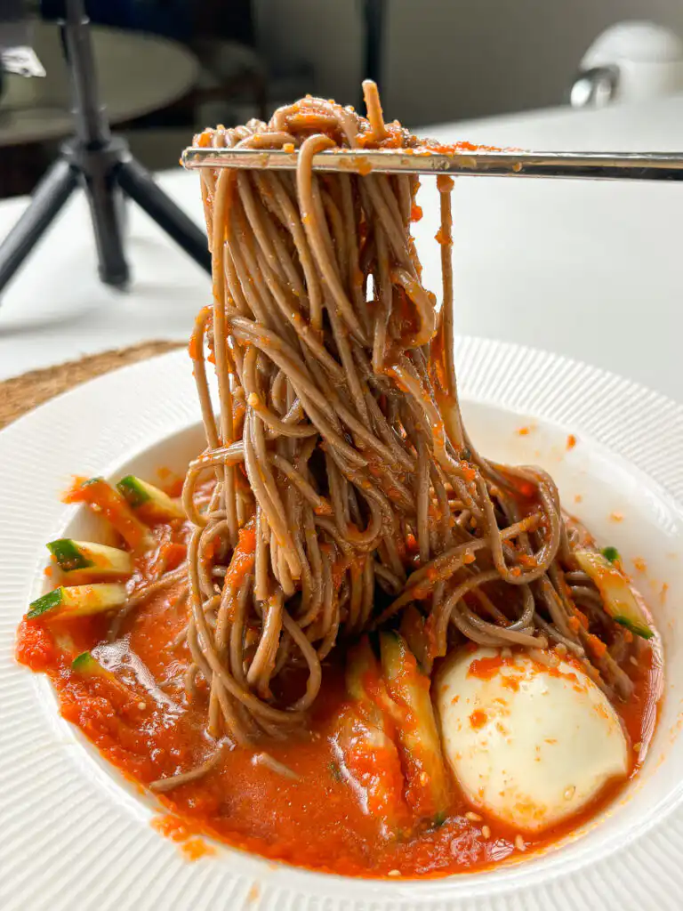 Tomato Gochujang Bibim Guksu - Summer Spicy Cold Noodles hand holding metal chopsticks lifting noodles from gochujang bibim guksu bowl