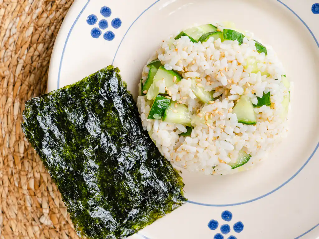 A bowl of low calorie kimbap rice mixture with diced cucumber and roasted sesame seeds next to seaweed sheets.