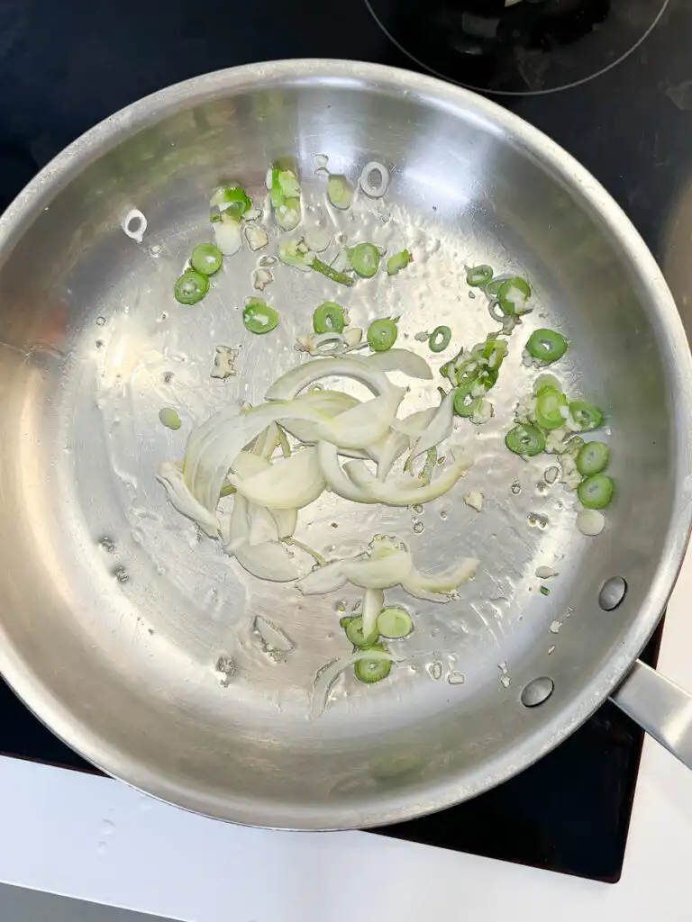 Adding sliced white onions to the pan with sautéed green onions and garlic.