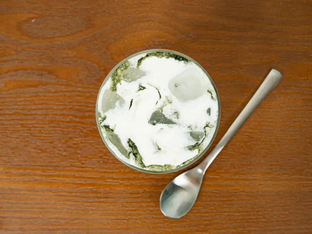 Korean Mugwort Latte: Easy Caffeine-Free Cream Ssuk Latte Overhead view of a teaspoon beside a glass of Korean Mugwort Latte, with mugwort powder visible on the cream foam